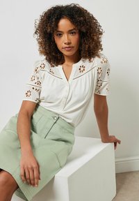 Camisa blanca con detalles de corte floral y botones, combinada con una falda verde clara. Cabello rizado, sentada sobre un bloque blanco. Fondo neutro.