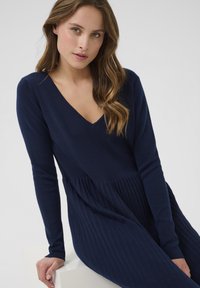 Jeune femme aux longs cheveux ondulés, portant une robe longue à manches longues et décolleté en V de couleur bleu marine, assise et reposant une main sur une surface blanche.
