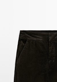 Czarne spodnie korduroy z pionowymi prążkami, z płaskim paskiem w talii i bocznymi kieszeniami. Materiał wydaje się miękki i teksturowany.