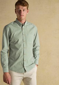 Zeleno-biele pruhované košele na gombíky s dlhými rukávmi, so vreckom na hrudi a hnedými gombíkmi, skombinované s svetlými nohavicami.