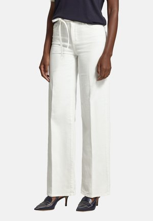 Donna che indossa pantaloni bianchi a vita alta e gamba larga con coulisse in vita, décolleté blu navy con punta a punta e una maglietta scura infilata dentro.