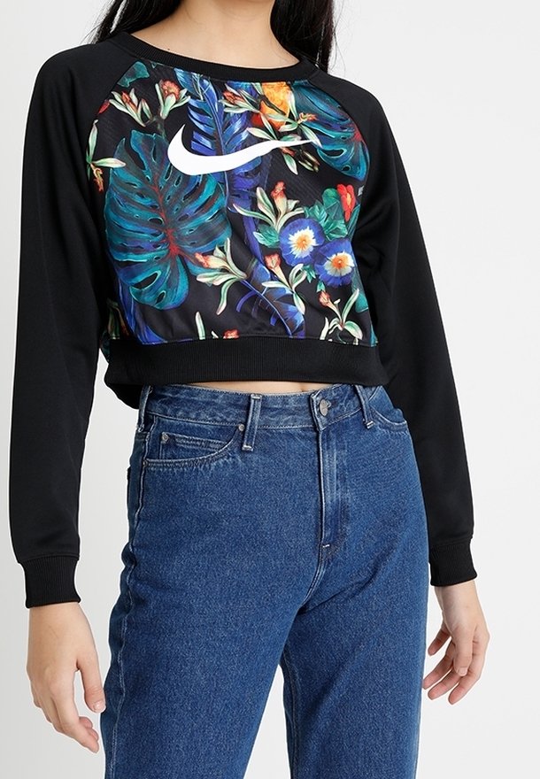 Svart kort sweatshirt med färgglad blommönster och Nike-logga på framsidan. Lång raglanärmar med ribbad mudd. Bärs med blå jeans.