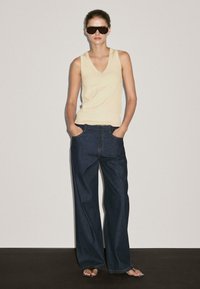Beige ærmeløs V-hals top sammen med mørkeblå brede denimbukser. Modellen har mørke solbriller på og står med hænderne i lommen.