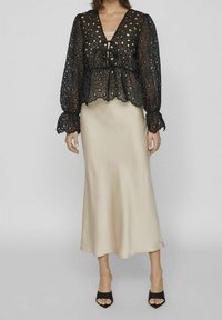Top in pizzo nero con ritagli floreali, maniche lunghe a palloncino e orlo scallop, abbinato a una gonna midi beige in satin e tacchi aperti neri.