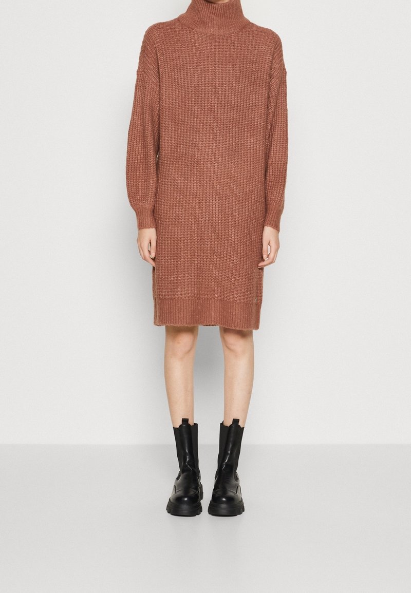 Personne portant une robe en maille col roulé couleur rouille jusqu'aux genoux, manches longues, et des bottines noires épaisses sur un fond uni.