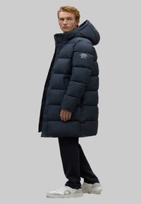 Abrigo acolchado azul marino con capucha, que presenta un diseño acolchado, puños elásticos y detalle de logo reflectante en el hombro. El modelo lleva pantalones negros y zapatillas.