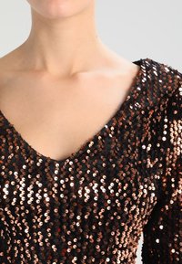 Svart topp täckt med kopparpaljetter, med en djup V-ringning och en figurnära form. Ytan verkar slät och reflekterande.