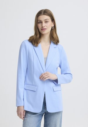 Kvinde med skulderlangt lyst brunt hår iført en lyseblå blazer over en grå top og falmede blå jeans, stående foran en ensfarvet hvid baggrund.