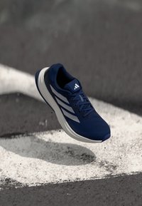 Zapatilla atlética azul con parte superior de malla, tres franjas blancas, suela acolchada y diseño con cordones. Diseñada para correr o para un uso activo.
