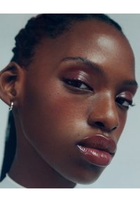 Lähikuva kasvoista, joissa on sileä, tumma iho. Kasvonpiirteisiin kuuluu rohkeat huulet, joissa on kiiltävä viimeistely, tummat silmät ja selkeät kulmakarvat. Yksinkertainen korvakoru koristeena.