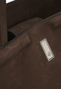 Bruine leren totebag met zachte textuur, voorzien van een zilveren logo plaque en contrasterende stiksels. Interieurzak zichtbaar.