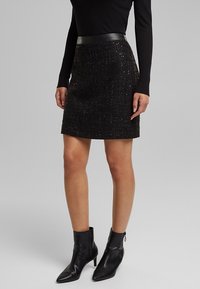 Mini gonna nera in tweed con finitura testurizzata e vita in similpelle nera liscia; abbinata a stivaletti neri alla caviglia con punta a punta.