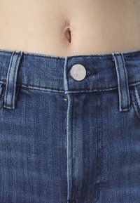 Tummansininen farkkuhousu, jossa on sopiva vyötärö, näkyvä hopea nappi ja keltainen ompelu. Kankaan pinnassa on lievää haalistumista ja sileä viimeistely.