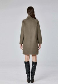 Manteau en laine kaki à la silhouette droite et aux manches longues, doté d'un col et d'une simple couture au dos. Associé à des bottes noires montantes.