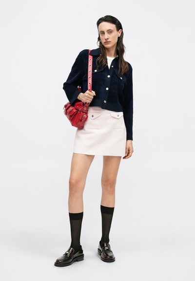 Mujer con una chaqueta corta de marine oscuro, falda mini rosa claro con bolsillos, medias altas negras hasta la rodilla, mocasines y llevando un bolso de hombro rojo.