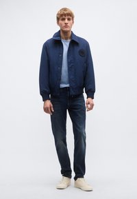 Μπουφάν bomber σε ναυτικό μπλε με υφασμένο λογότυπο, συνδυασμένο με ανοιχτό μπλε μπλουζάκι με στρογγυλή λαιμόκοψη, σκουρόχρωμο τζιν και μπεζ αθλητικά παπούτσια.