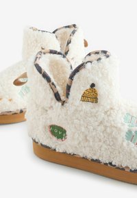 Karvased valged sussid tekstuuriga pinnaga, millel on värvikas tikand, sealhulgas kollane müts, "LOVE" silt ja mustriline serv.