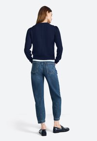 Marineblauer Strickpullover mit Puffärmeln und strukturiertem Rücken, kombiniert mit hochgeschnittenen, verblassten blauen Jeans und schwarzen Slippern.