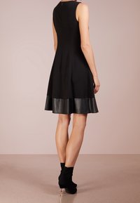 Robe noire sans manches avec un corsage ajusté et un ourlet évasé bordé de cuir. Tissu lisse avec une texture mate, fermeture éclaire au dos.