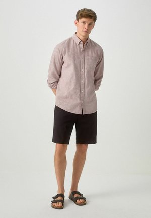 Junger Mann steht mit hinter dem Rücken verschränkten Händen, trägt ein hellrosa Hemd mit Knöpfen, schwarze Shorts und schwarze Sandalen vor schlichtem Hintergrund.