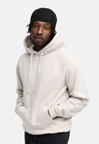 Hellgrau Kapuzenpullover mit einer Fronttasche, Kordelzügen und gefütterter Kapuze. Getragen mit schwarzen Hosen. Der Stoff sieht weich und bequem aus.