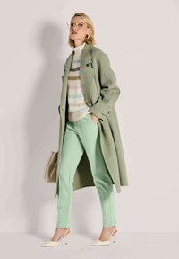 Vrouw in een pastelgroene jas en broek, gestreepte coltrui, beige hakken, en een beige handtas, tegen een effen achtergrond.