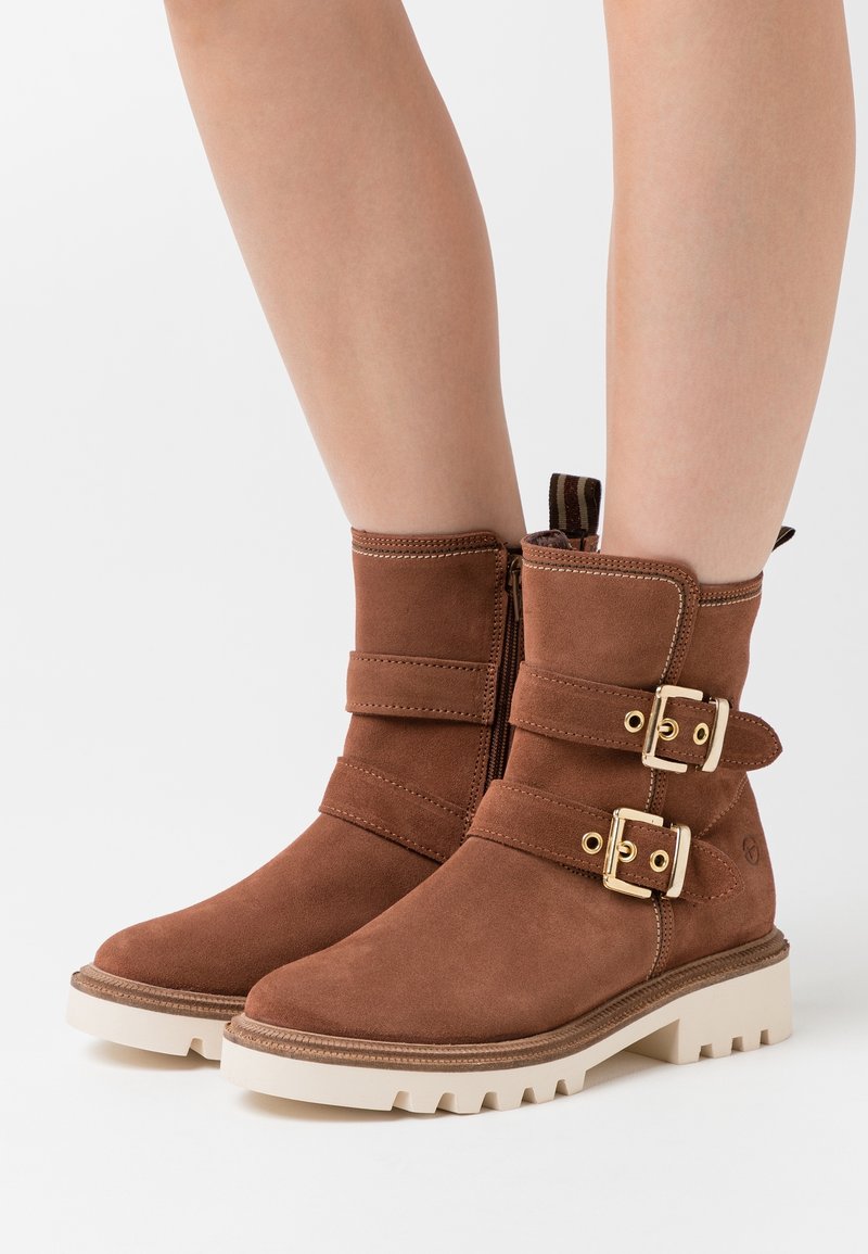 Bottines en daim marron avec double boucle dorée, fermeture éclair latérale et semelles épaisses blanches à crampons, portées sur jambes nues.