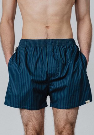 Torse masculin portant un short rayé bleu foncé avec taille élastique et poches, mains dans les poches, debout devant un fond uni.