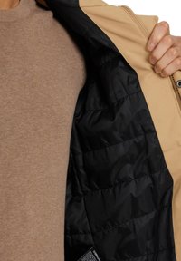 Beige ytterjacka med svart quiltad foder. Har dragkedja och en etikett längst ner. Beigetröja synlig under.