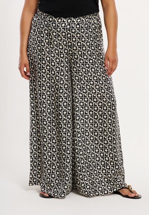 Person iført vidbenede sorte og hvide ternede bukser med cirkulære mønstre, sort top og sorte sandaler med gulddetalje.