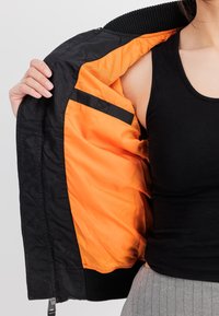 Svart bomberjakke laget av quiltet stoff, med oransje fôr, ribbet krage og en sidelomme med en svart stropp.