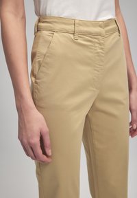 Pantaloni beige con una texture liscia, dotati di tasche laterali e una vestibilità sartoriale. Design a vita alta, arricchito da dettagli di cucitura sottili.