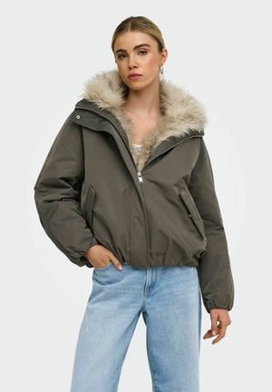 Jeune femme portant une veste d'hiver vert olive avec capuche doublée de fourrure et un jean bleu clair, debout avec une main dans la poche devant un fond uni.