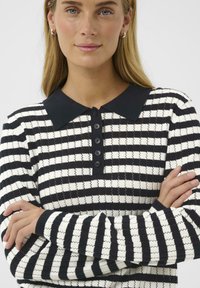 Una donna con capelli biondi lisci indossa una camicia a maniche lunghe in bianco e nero a scacchi con colletto e bottoni neri, con le braccia incrociate.