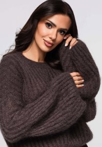 Joven con cabello largo y oscuro que lleva un suéter acanalado de color marrón oscuro y borroso, sosteniendo suavemente la manga cerca de su rostro y sonriendo.