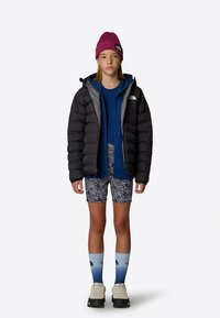 Mladić u crnoj puffer jakni, plavoj dukserici, šarenim šortsevima, čarapama s plavim gradijentom, bijelim cipelama i crvenom kapom stoji naslonjen na bijelu pozadinu.