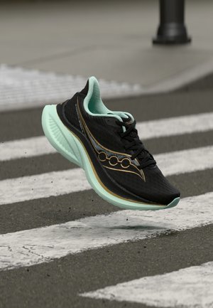 Schwarzer und mintgrüner Laufschuh mit dicker Sohle in der Luft über einem Zebrastreifen auf einer Asphaltstraße.