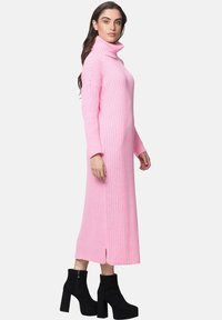 Langes, pinkes, geripptes Kleid mit hohem Rollkragen und seitlichem Schlitz, aus weichem Stoff, kombiniert mit schwarzen, schulterhohen Stiefeln mit Absatz.
