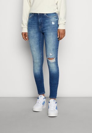 Vrouw draagt lichtgewassen skinny jeans met gescheurde details en witte sneakers met blauwe strepen, staand tegen een effen achtergrond.