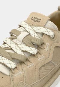 Les baskets en daim beige présentent des accents texturés, des lacets crème à motif et un logo sur la languette. Le design comprend des détails cousus et un rembourrage.