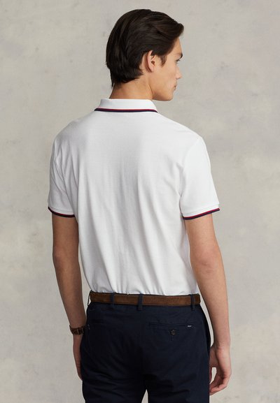 Polo branco com detalhes em vermelho e azul-marinho na gola e nas mangas, combinado com calças azul-marinho. Cinto castanho visível e bolso traseiro com botão.
