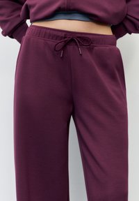 Bordowe spodnie dresowe wykonane z miękkiego materiału, z sznurkiem w talii, luźnym krojem i bocznymi kieszeniami. Gładka tekstura bez widocznych wzorów.
