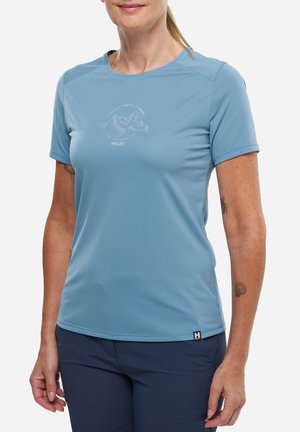 Femme portant un t-shirt bleu clair à manches courtes avec un logo de montagne et un pantalon bleu marine, debout devant un fond blanc uni.