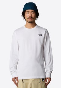 Witte longsleeve t-shirt van katoen met een klein zwart logo op de borst. De model draagt een blauwe gebreide beanie.