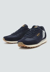 Pár námořnických modrých tenisek Patrick Monza s bílými pruhy po stranách a gumovými podrážkami na světle šedém pozadí.