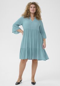 A loose, knee-length dress in light teal fabric, featuring a V-neck, three-quarter bell sleeves, and a tiered, ruffled hem.