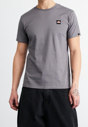 Personne portant un t-shirt gris uni à manches courtes avec un petit logo sur la poitrine, un pantalon noir ample et des tatouages visibles sur les deux bras et la main gauche.