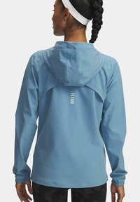 Mujer con el cabello trenzado que lleva una diadema blanca y una chaqueta con capucha de color azul claro con tiras reflectantes en la parte posterior, de pie frente a un fondo liso.