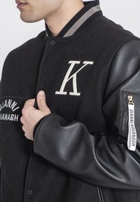 Čierna bunda z vlnenej zmesi s koženými rukávmi, ktorá má veľkú nášivku "K" a štítok GIANNI KAVANAGH na ľavom rukáve. Pruhovaný golier.