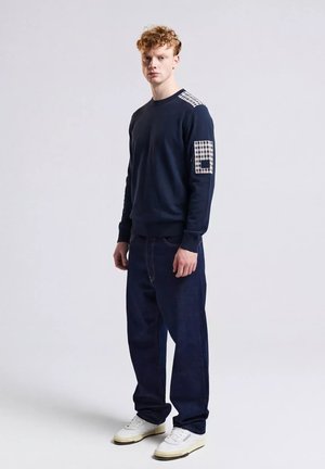 Giovane uomo con capelli ricci che indossa un maglione blu navy con toppe a scacchi e jeans scuri a gamba larga, in piedi contro uno sfondo chiaro e uniforme.